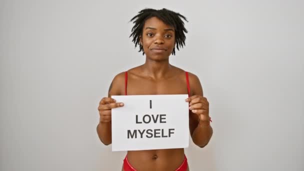 Vreugdevolle jonge zwarte vrouw met dreadlocks, vol vertrouwen met 'love my body' teken, pronkend met haar tandenglimlach, straalt positiviteit uit terwijl ze geïsoleerd staat tegen een witte achtergrond. - Video