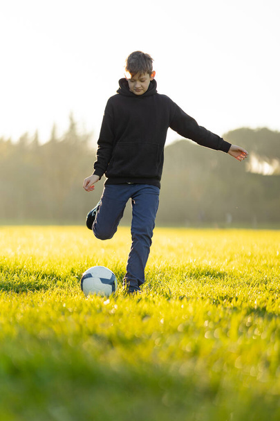 Een jongen schopt een voetbal in een veld. De jongen draagt een zwarte hoodie en een blauwe jeans. - Foto, afbeelding