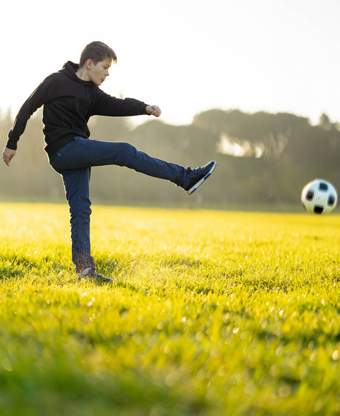 Un chico patea una pelota de fútbol en un campo. El chico lleva una sudadera con capucha negra y vaqueros azules. - Foto, imagen