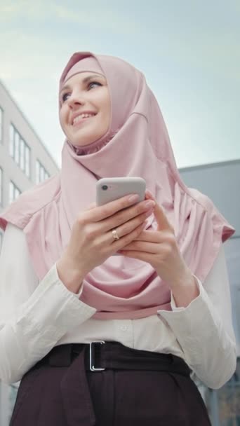 Pionowy ekran: Piękna muzułmanka stojąca w pobliżu nowoczesnego biurowca ze smartfonem w rękach. Muzułmańska bizneswoman z komórką stojąca w pobliżu centrum biurowego. Dziewczyna z telefonem uśmiechnięta. - Materiał filmowy, wideo