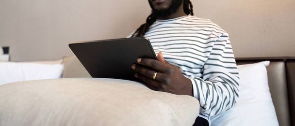 Uśmiechnięty młody Afroamerykanin korzystający z tabletu cyfrowego, siedzący na łóżku w domu. koncepcja komunikacji technologicznej online. - Zdjęcie, obraz