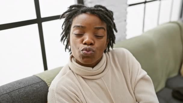 Selbstbewusst und liebenswert sitzt eine junge schwarze Frau mit Dreadlocks gemütlich auf dem Sofa und pustet einen verspielten Kuss in die Kamera. Sexy und schöne Liebe ausdrücken, zu Hause. - Filmmaterial, Video