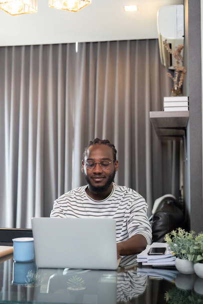 Afrikkalainen amerikkalainen mies työskentelee kannettavan tietokoneen kaukosäätimen kanssa istuessaan lasipöydässä olohuoneessa. Musta kaveri tekee freelance työtä kotitoimistossa. - Valokuva, kuva