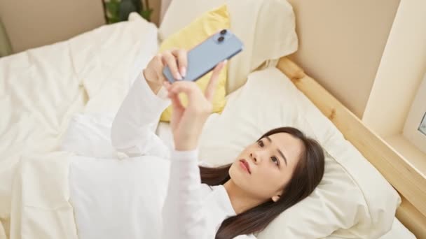 Μια νεαρή Ασιάτισσα ξαπλώνει στο κρεβάτι βγάζοντας μια σέλφι με το smartphone της σε ένα φωτεινό υπνοδωμάτιο.. - Πλάνα, βίντεο