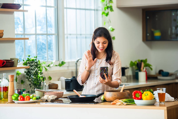India asiático joven mujer usando smartphone en cocina mientras cocinar - Foto, imagen