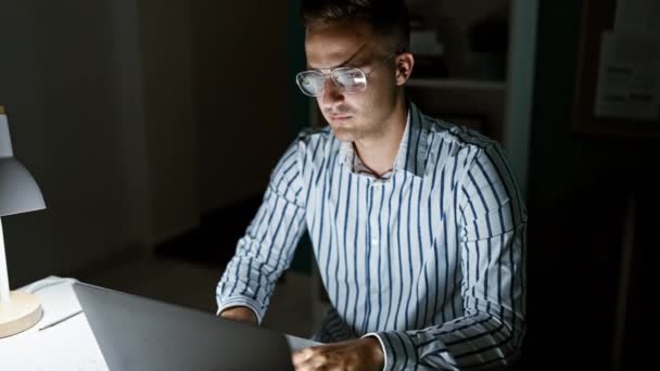 Un uomo concentrato che lavora fino a tardi sul suo portatile in una stanza buia dell'ufficio, illuminato da una lampada da scrivania. - Filmati, video