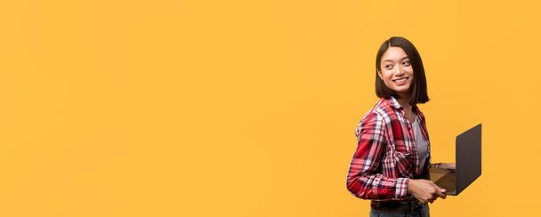 Nuori aasialainen nainen ruudullisessa paidassa pitää iloisesti kannettavaa tietokonetta, jossa on runsaasti kopiotilaa eloisalla keltaisella taustalla. - Valokuva, kuva