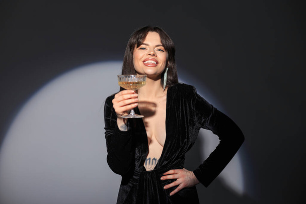 Veselá krásná okouzlující módní žena s úsměvem v módní černé sexy šaty s výstřihem drží sklenici a slaví ve studiu na tmavém pozadí - Fotografie, Obrázek