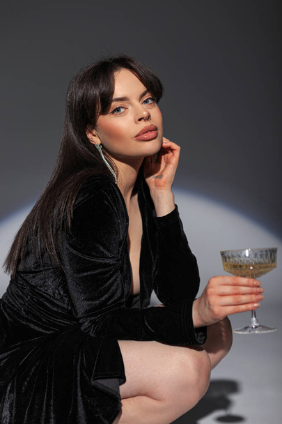 Όμορφη λαμπερή νεαρή γυναίκα σε ένα μοντέρνο μαύρο σέξι φόρεμα κρατά ένα ποτήρι σαμπάνια που κάθεται και ποζάρει στο στούντιο σε ένα σκοτεινό φόντο. Αρκετά κομψή κυρία γιορτάζει τις διακοπές - Φωτογραφία, εικόνα