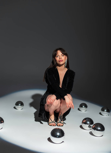 Сексуальная гламурная модель в стильном сексуальном платье с декольте на каблуках сидит на полу с зеркальными шариками в студии на темном фоне - Фото, изображение