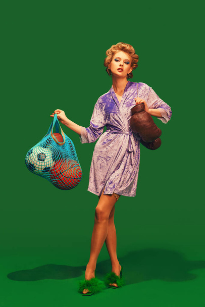 Mujer joven en albornoz sedoso con rizadores de pelo con guantes de boxeo y pelotas deportivas sobre un fondo verde vibrante. Concepto de deporte, competición, estilo de vida activo, hobby, recreación. Anuncio - Foto, Imagen