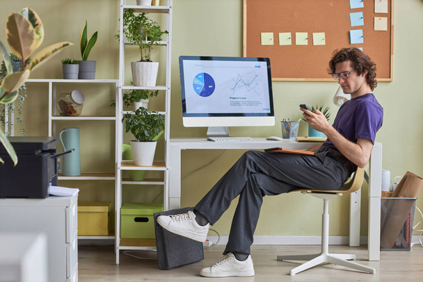 Pełna długość widok z boku portret młody człowiek relaks w miejscu pracy w biurze i za pomocą smartfona noszenia ubrań casual, przestrzeń kopiowania - Zdjęcie, obraz