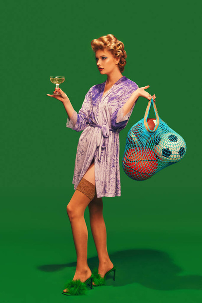 Mujer joven en albornoz sedoso con rizadores para el cabello sosteniendo un vaso de bolas deportivas de cóctel sobre un fondo verde vibrante. Concepto de deporte, competición, estilo de vida activo, hobby, recreación. Anuncio - Foto, imagen
