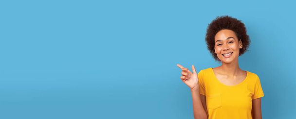Sarı bluzlu, gülümseyen Afro-Amerikan kadın mavi arka plandaki boşluğu işaret ederek, internete bağlanan bir afiş ya da maket yapıyor. - Fotoğraf, Görsel