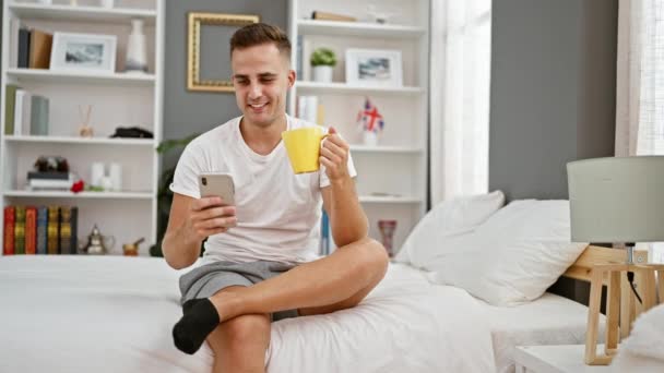 Uomo sorridente che tiene smartphone e tazza di caffè si siede sul letto in camera da letto ben arredata con arredamento bianco e grigio. - Filmati, video