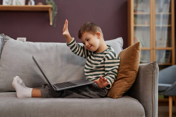 Seitenansicht Porträt eines glücklichen kleinen Jungen mit Down-Syndrom mit Laptop auf dem Sofa und winkt zum Videochat, Kopierraum - Foto, Bild