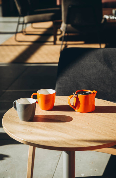 Twee kopjes en een theepot op een tafel in een café met harde schaduwen. Oranje theepot en kopjes in het interieur van een modern café. Zonnige ochtend en een kop thee. Kopieerruimte. - Foto, afbeelding