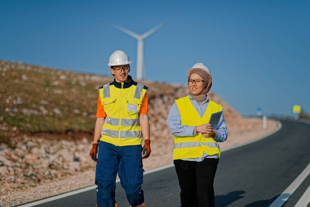 Una ingeniera con una tableta y su colega inspeccionan turbinas eólicas en un campo, demostrando trabajo en equipo y dedicación a proyectos de energía renovable.  - Foto, Imagen