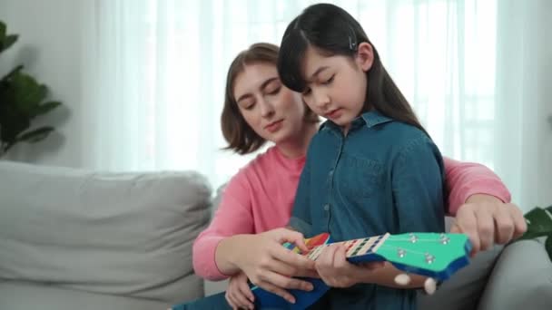 Szczęśliwa dziewczyna gra ukulele podczas gdy kaukaska mama uczy i wyjaśnić o akustycznej muzyki w domu. Słodkie dziecko uczy się o instrumencie. Szczęśliwa, biała matka i dziewczyna spędzają razem czas. Pedagogika. - Materiał filmowy, wideo