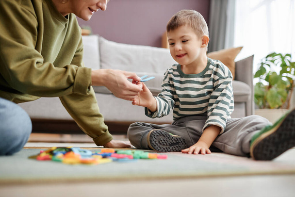 Porträt eines glücklichen kleinen Jungen mit Down-Syndrom, der mit Buchstaben auf dem Boden spielt und Spaß am Lernen mit Mama hat - Foto, Bild