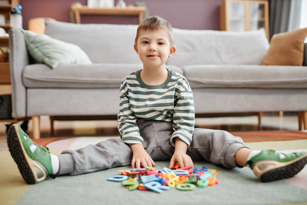 Retrato de comprimento total de menino com síndrome de down brincando com brinquedos sentados no chão e olhando para a câmera - Foto, Imagem