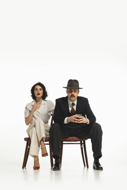 Элегантная стильная пара, мужчина и женщина в классических костюмах сидят на стуле с эмоциональным лицом и смотрят шоу, телевизор изолирован на белом фоне. Концепция ретро и винтажа, мода, отношения - Фото, изображение