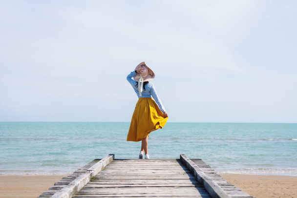 Dívka v klobouku a žluté sukni stojí na dřevěném molu na pozadí moře. Odpočiň si u moře. - Fotografie, Obrázek