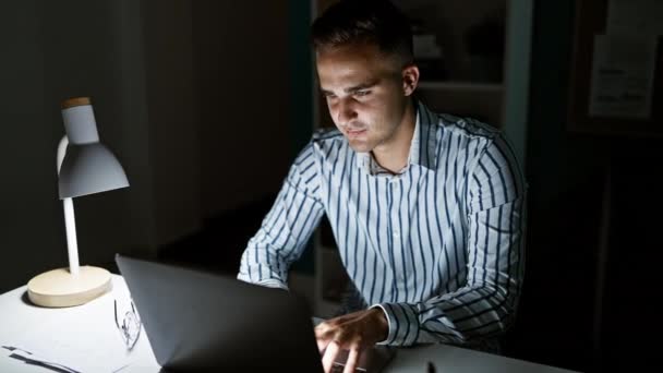 Un joven hispano enfocado trabajando hasta tarde en la oficina, iluminado por la luz de la lámpara de escritorio. - Metraje, vídeo