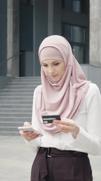 垂直スクリーン: 正式な服やヒジャーブで美しいイスラム教徒の女性は,クレジットカードとスマートフォンでオンラインショッピングを行います. オフィスビルの近くに立って簡単な支払いを楽しんでいる若い女性 - 映像、動画