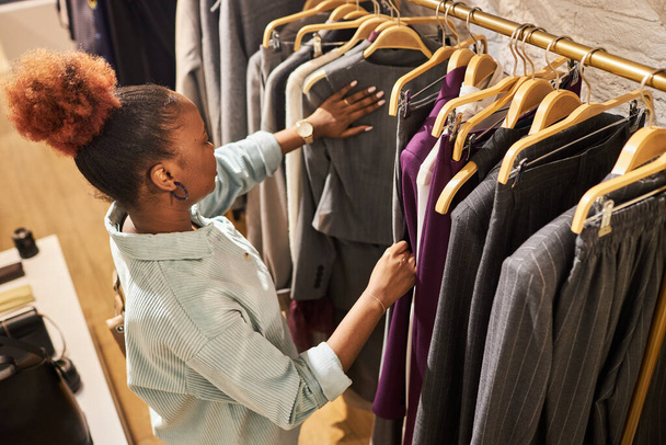 Retrato de ángulo alto de la joven afroamericana que navega por la ropa en el estante en la tienda de ropa, espacio de copia - Foto, imagen