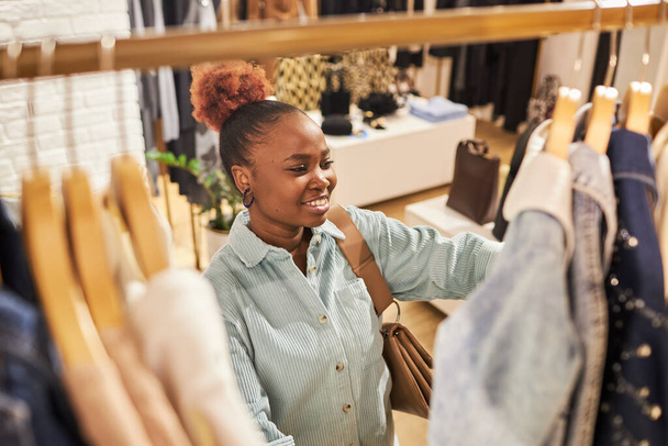 Retrato de alto ángulo de mujer negra sonriente buscando ropa en el estante en la tienda de ropa y disfrutando de compras, espacio para copiar - Foto, Imagen
