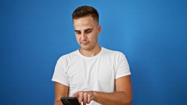 Молодий дорослий чоловік взаємодіє зі смартфоном на ізольованому синьому фоні, демонструючи сучасне спілкування. - Кадри, відео