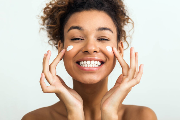 Una alegre dama afroamericana con una sonrisa brillante aplica crema facial, simbolizando el cuidado de la piel y las rutinas de cuidado personal - Foto, Imagen