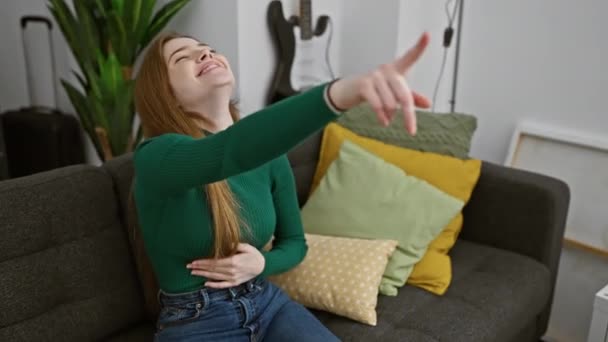 Cheerful young blonde woman at home wearing a sweater, pointing a 'gotcha' finger at you, doubling over with infectious laughter and teasing joy! - Footage, Video