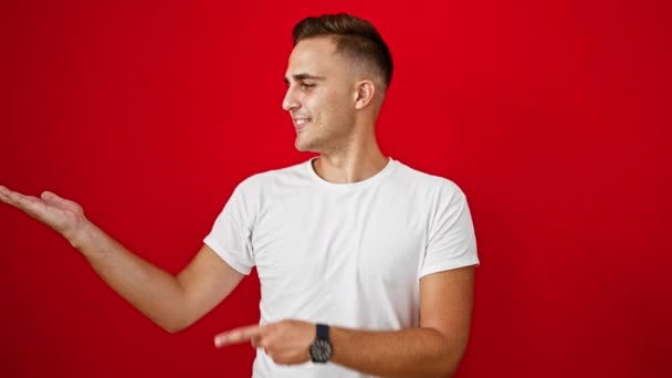 Beyaz tişörtlü gülümseyen İspanyol genç adam izole edilmiş kırmızı arka planda el hareketleriyle. - Video, Çekim