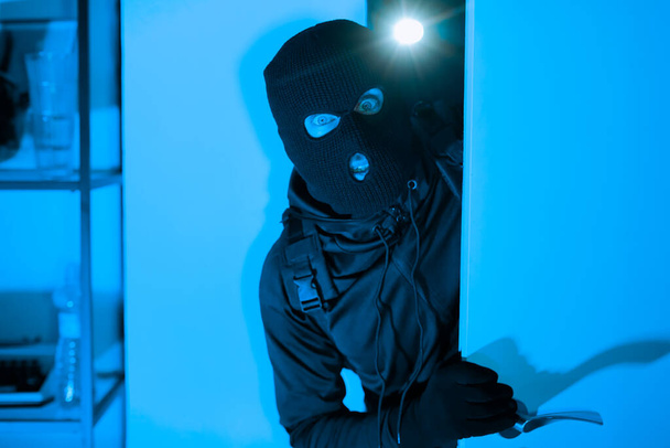 Un ladro mascherato con un piede di porco sta entrando in una stanza con l'intento di rubare, catturato in una fioca luce blu suggerendo attività criminali notturne - Foto, immagini