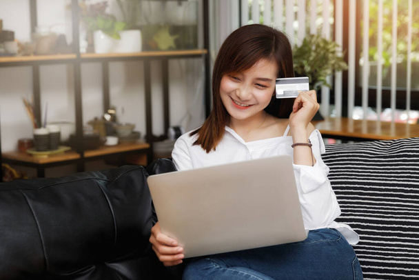 Щаслива молода азіатка тримає кредитну картку і ноутбук, сидячи на дивані вдома
. - Фото, зображення