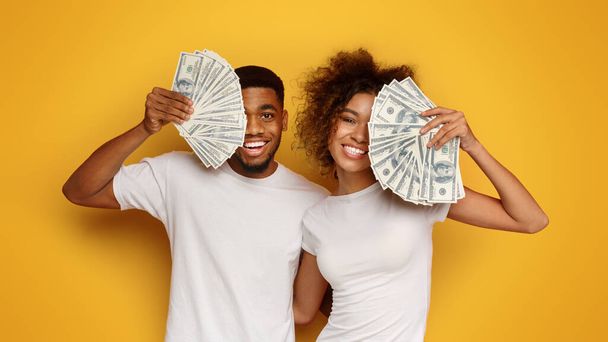 Счастливая пара. Радостные афро-американец и женщина покрывают лица с долларовыми фанатами, оранжевый фон студии - Фото, изображение