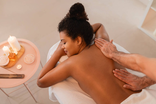 Vista de ángulo alto de la mujer afroamericana acostada en la mesa de masaje con velas alrededor disfrutando de un masaje de espalda - Foto, imagen