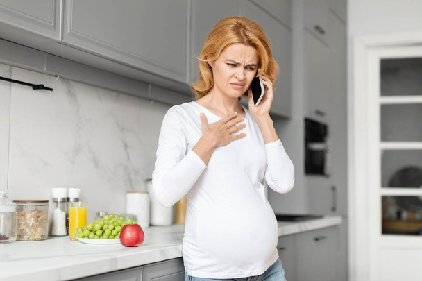 Zaniepokojona Europejka w ciąży czuje dyskomfort w kuchni podczas rozmowy telefonicznej, wskazując na znaczenie odżywiania, dzwoniąc do lekarza - Zdjęcie, obraz