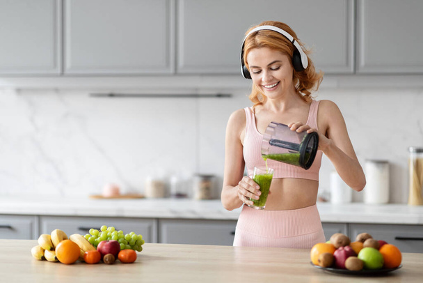 Una mujer alegre en traje de fitness está vertiendo un batido verde de una licuadora en un vaso en una cocina moderna con frutas en la mesa - Foto, Imagen