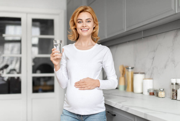 Una donna incinta europea radiosa nella sua cucina con un bicchiere d'acqua, che simboleggia una sana idratazione e nutrizione prenatale. - Foto, immagini