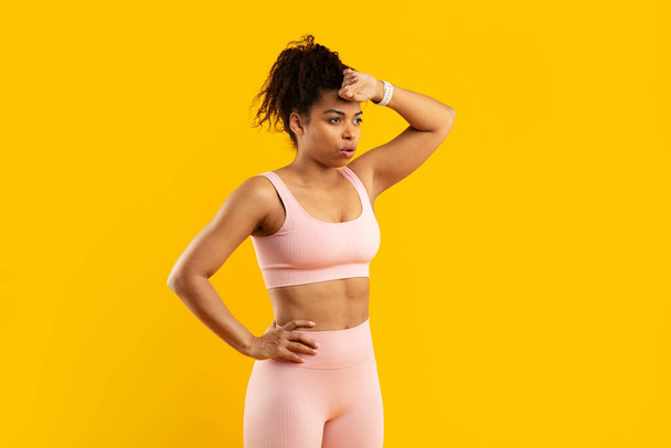Dieses lebendige Bild zeigt eine afrikanisch-amerikanische Dame in Fitnesskleidung, die sich vor einem isolierten gelben Hintergrund den Schweiß von der Stirn wischt. - Foto, Bild