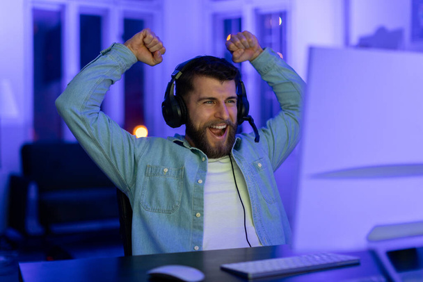 Een millennial gamer vreugdevol viert een overwinning in zijn gaming setup thuis, belichaamt de euforie en verslaving van gaming cultuur - Foto, afbeelding