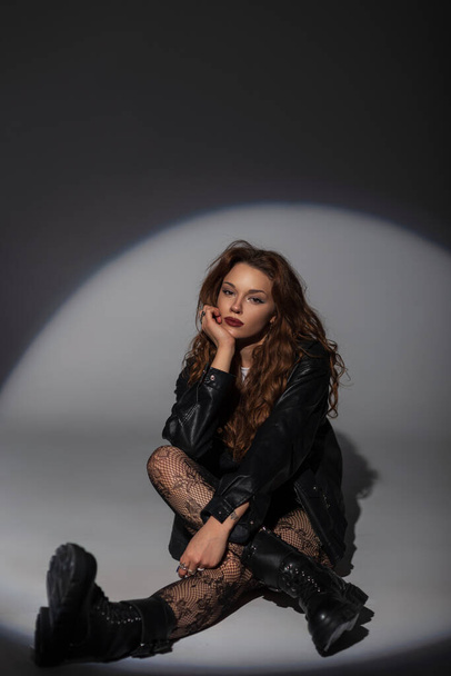 Stylish beautiful glamorous sexy woman in a fashion black leather jacket with lace tights and boots sits on the floor on a dark background in the studio - Photo, Image