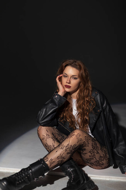 Élégante belle mode rock femme sexy dans une veste en cuir noir à la mode avec des collants et des bottes en dentelle assis et posant sur le sol dans le studio sur un fond sombre - Photo, image