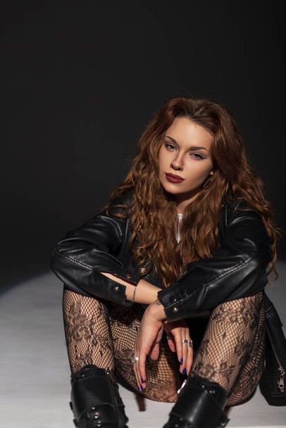 Stylish beautiful fashion sexy woman in a fashionable black rock leather jacket with lace tights sits and poses on a dark background - Photo, Image