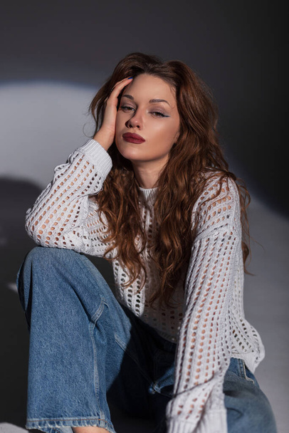 Moda fresca bella ragazza modello in un maglione bianco alla moda con jeans siede sul pavimento in studio su uno sfondo scuro. Chic signora - Foto, immagini