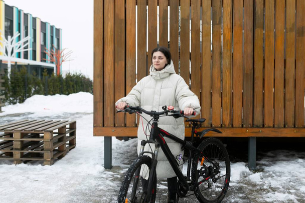 Een vrouw staat naast een fiets in een besneeuwd landschap. Ze draagt winterkleding en lijkt de fiets te onderzoeken.. - Foto, afbeelding