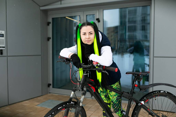 Μια γυναίκα με μαύρη και πράσινη στολή στέκεται δίπλα σε ένα ποδήλατο. Είναι σε ένα αστικό περιβάλλον, αναζητούν αυτοπεποίθηση και κομψό. - Φωτογραφία, εικόνα